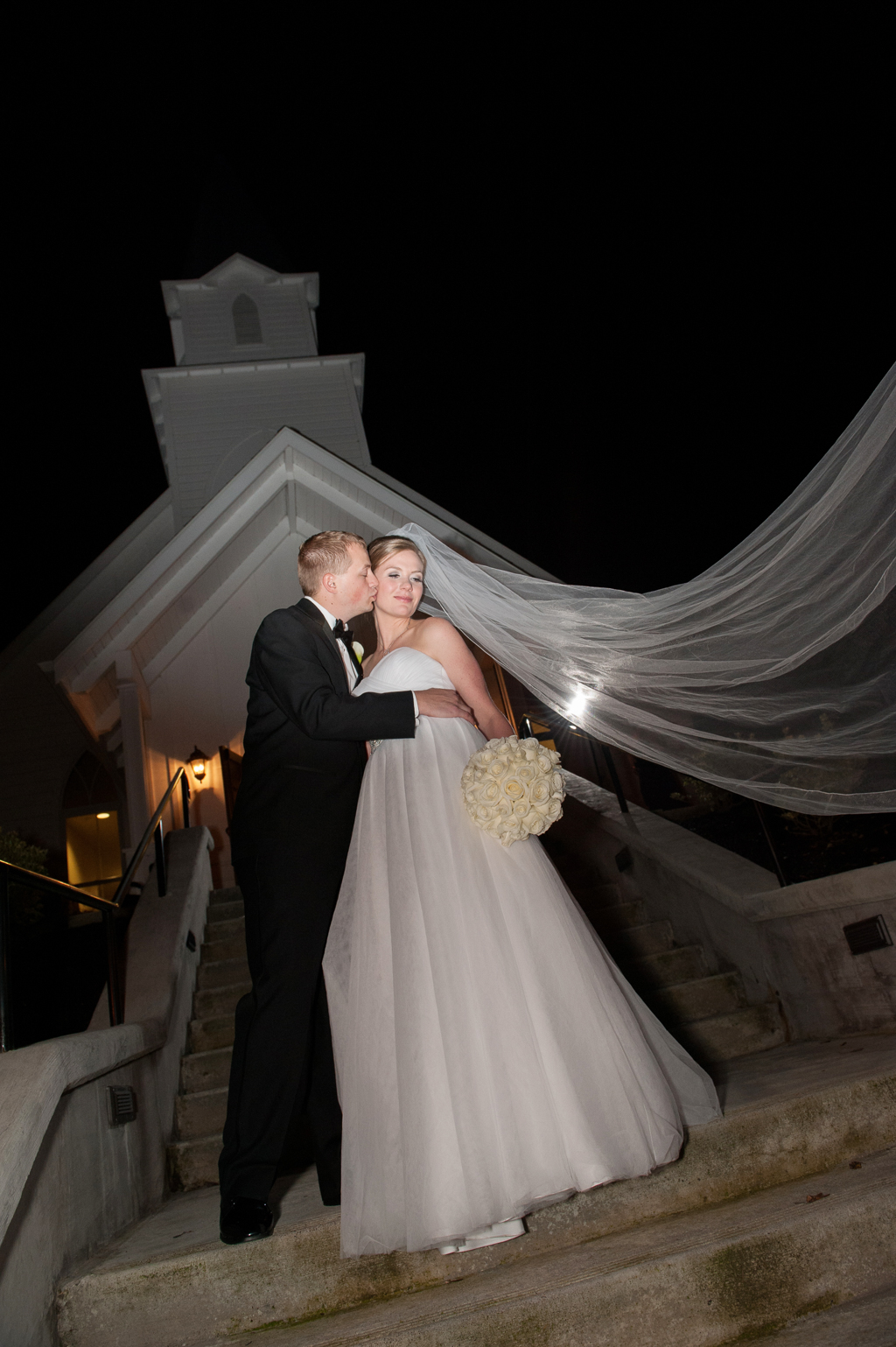 groom kisses bride in front of abernathy chapel and brides long veil flies in wind
