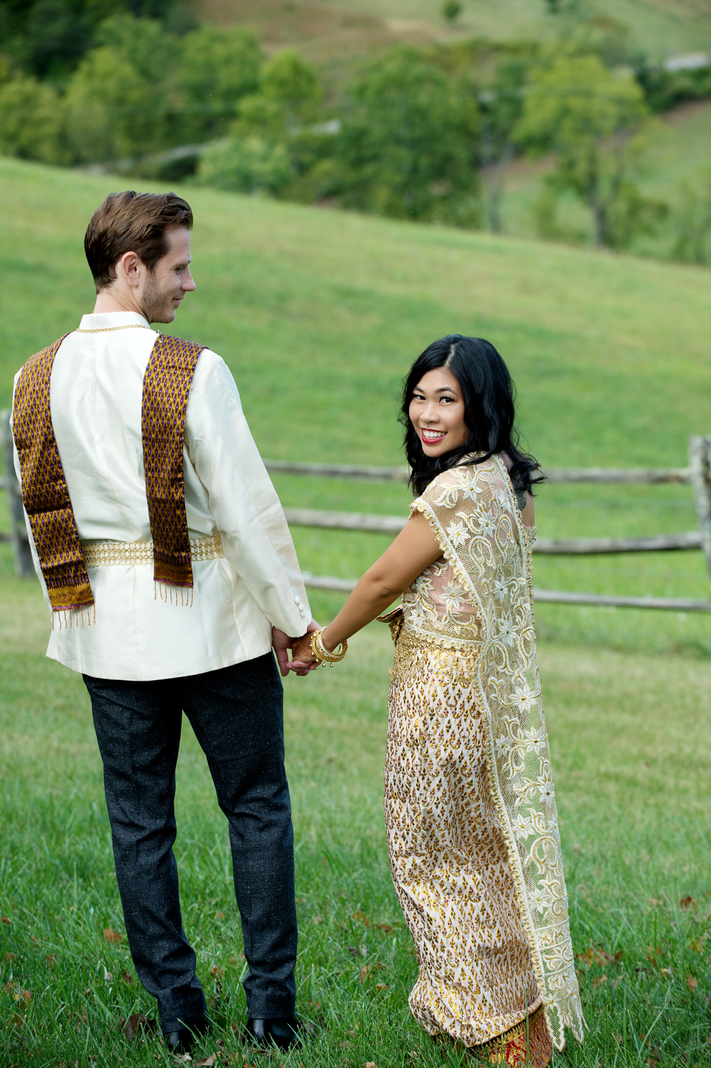 a couple in traditional gold cambodian wedding attire walk hand in hand down a rolling pasture