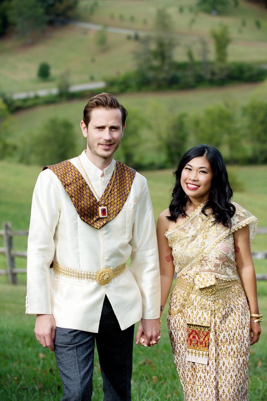 a couple in traditional gold cambodian wedding attire walk hand in hand up a rolling pasture