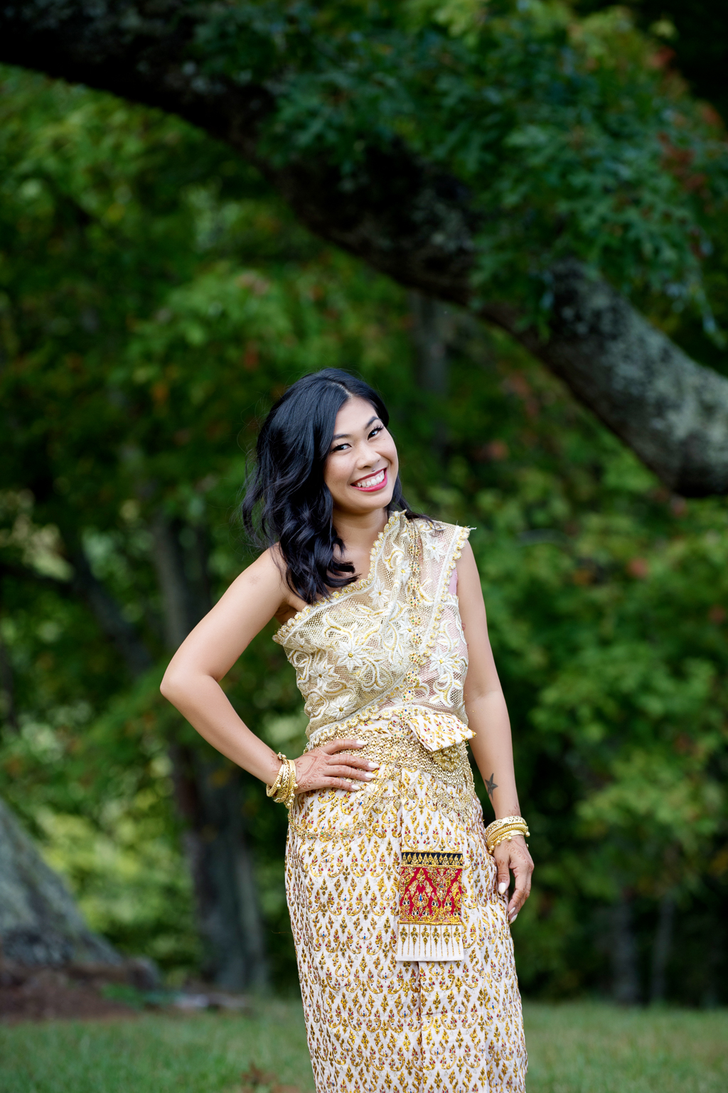 a woman in traditional gold cambodian wedding attire smiles at the camera in front of a dark green tree