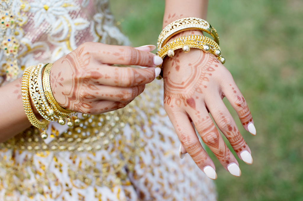 a woman with traditional cambodian gold wedding attire and jewelry fixes a bracelet on her hand that is covered in henna