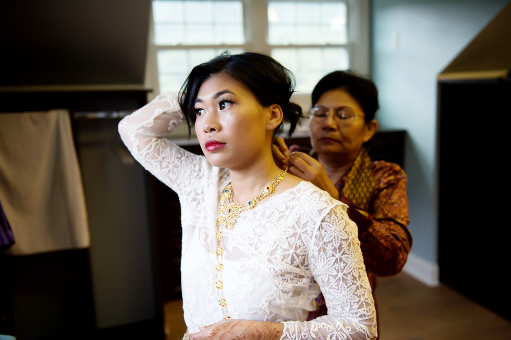 a mother helps a bride in a white lace dress with her traditional cambodian jewelry
