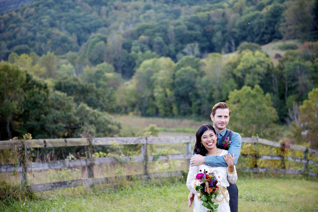 a bride carrying colorful wildflowers and wearing a white lace dress and traditional cambodian jewelry hug in a field in front of the blue ridge mountains