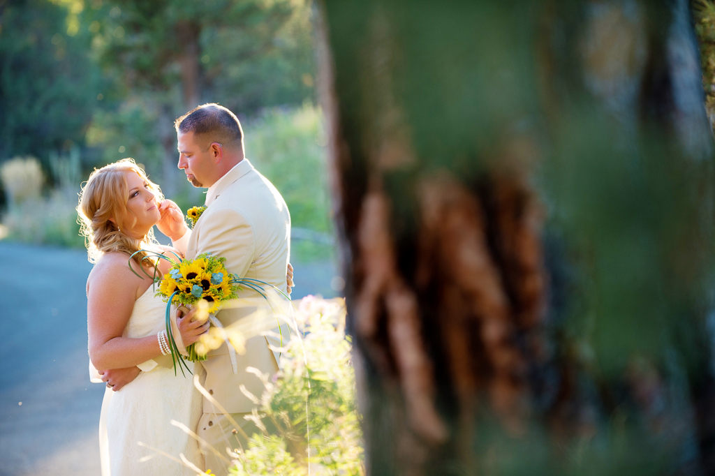 a bride holds a sunflower bouquet as her groom hugs her in the sunshine behind a tree
