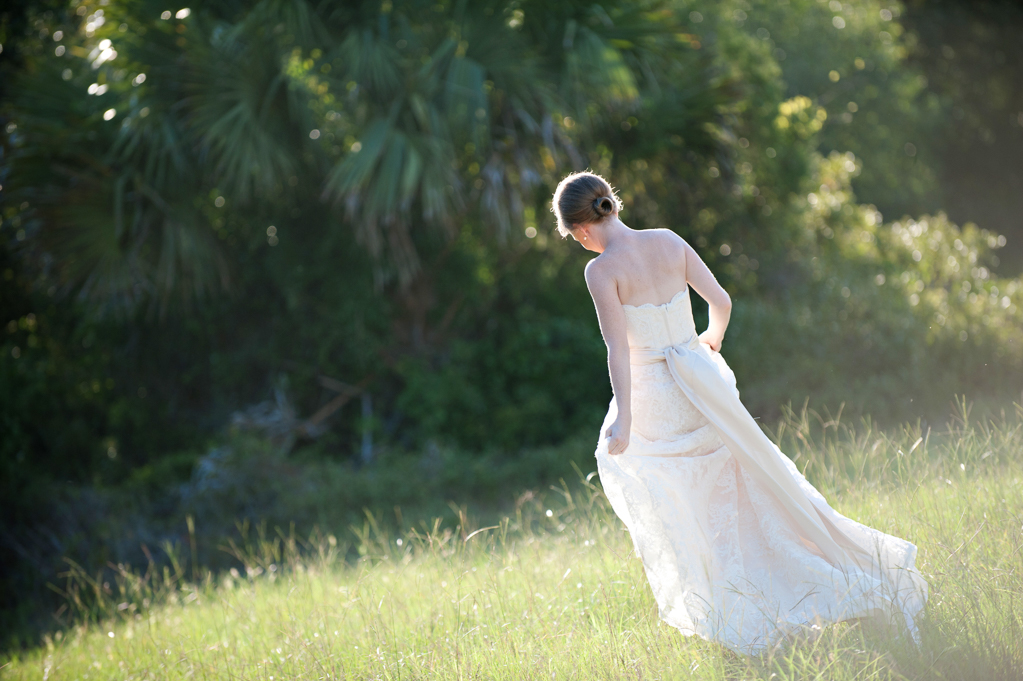 a bride walks through a sunlit field holding her wedding dress