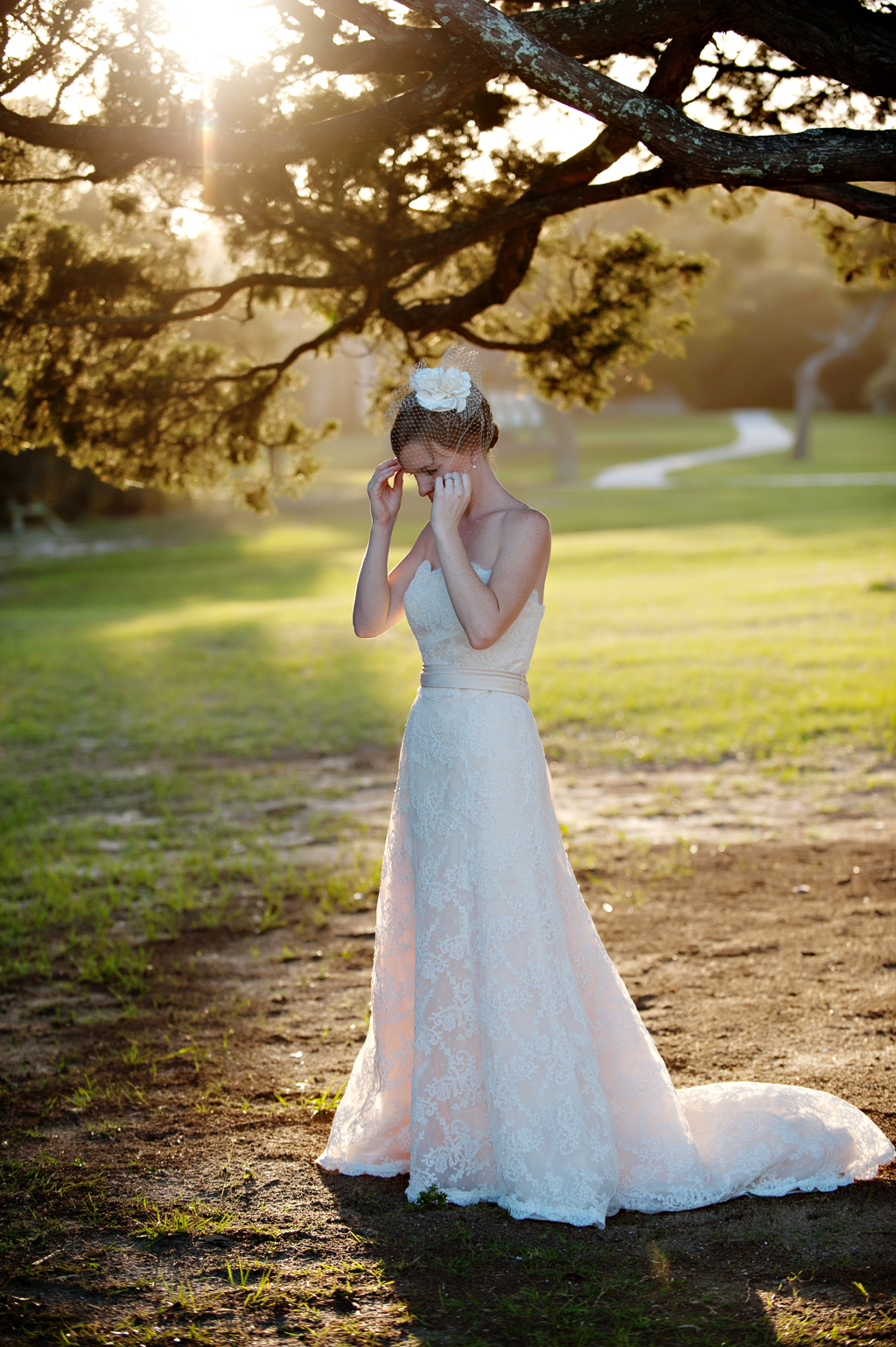 a bride bathed in sunlight under an oak tree fixes her birdcage veil