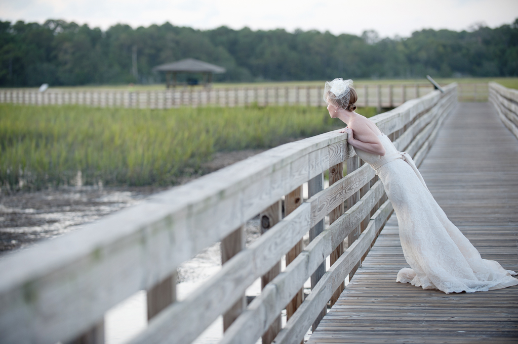 a bride wearing a birdcage veil leans over a boardwalk over murrells inlet looking at the marsh sound