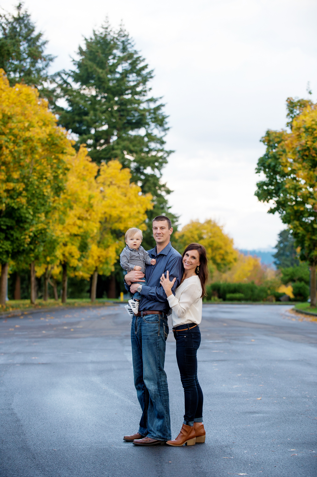 a mom and dad holding a baby boy stand in the middle of a street lined with yellow autumn trees