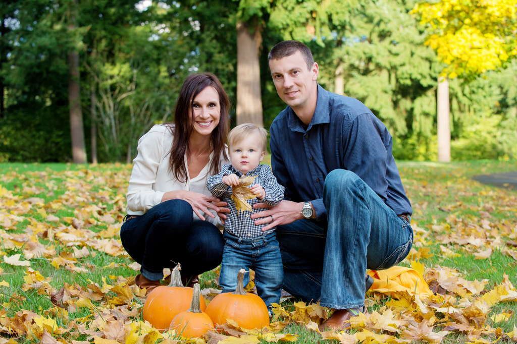 a mom and dad kneel around a baby boy playing with yellow leaves and pumpkins