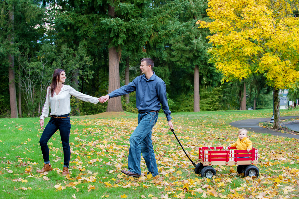 a mom pulls dad by the hand through a yard filled with yellow leaves as dad pulls a baby boy in a red wagon