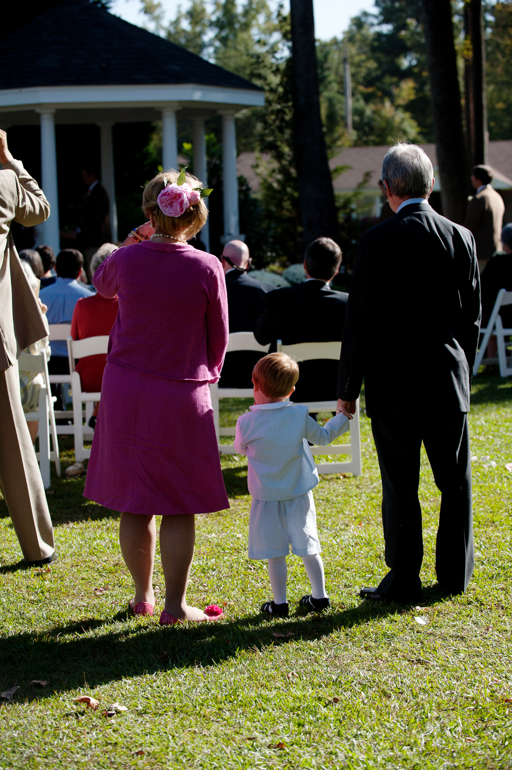 a little boy in a seersucker suit holds his grandpas hand looking on to a wedding ceremony