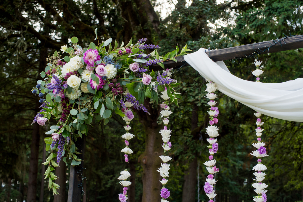 strings of purple and ivory flowers hang from a wedding ceremony arbor