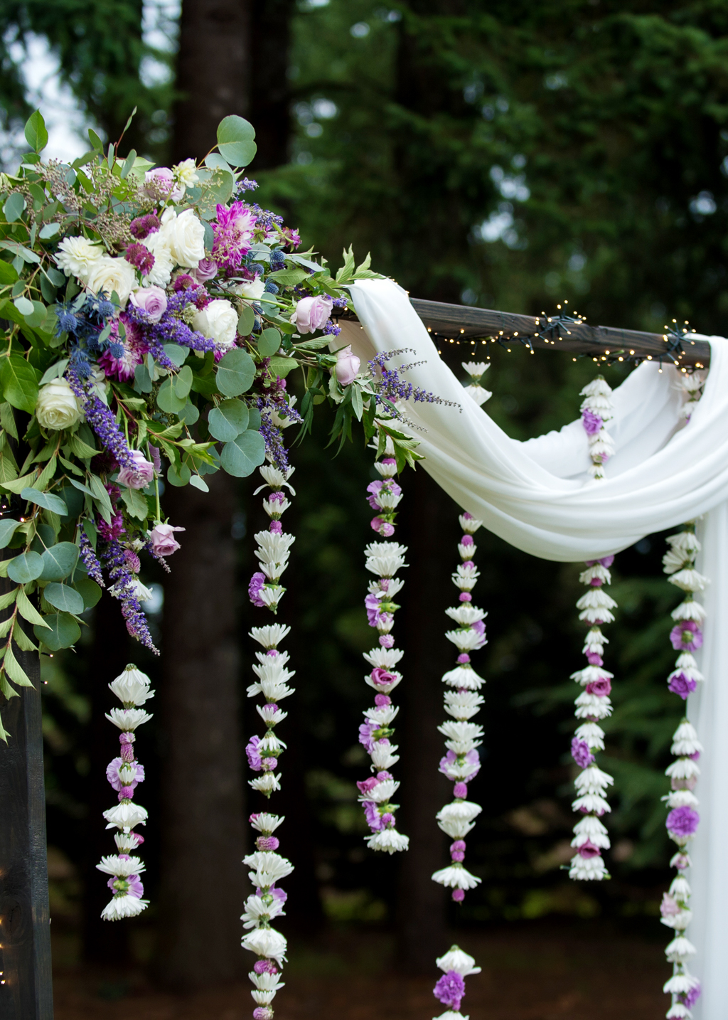 strings of purple and ivory flowers hang from a wedding ceremony arbor draped with white fabric