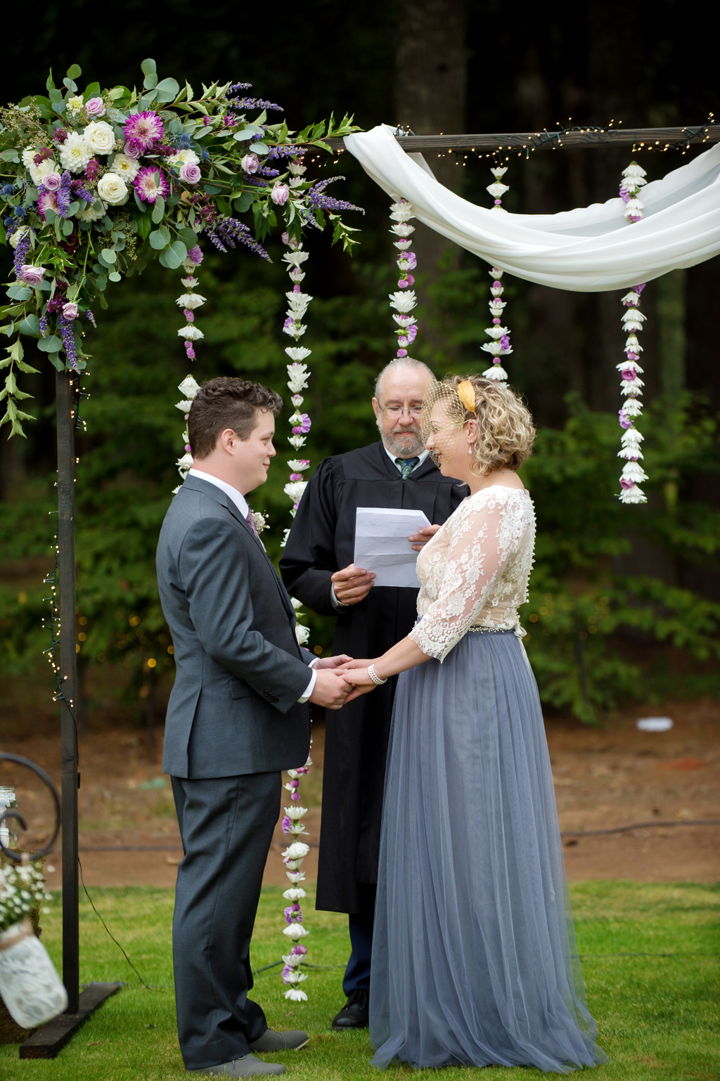 a bride and groom stand in front of a wedding arbor strung with white and ivory flowers