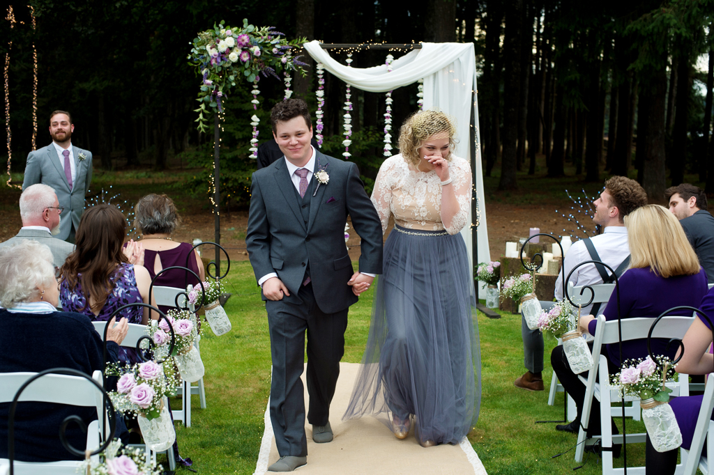 a bride wearing a flowy gray skirt and ivory lace top walks with her groom down the aisle