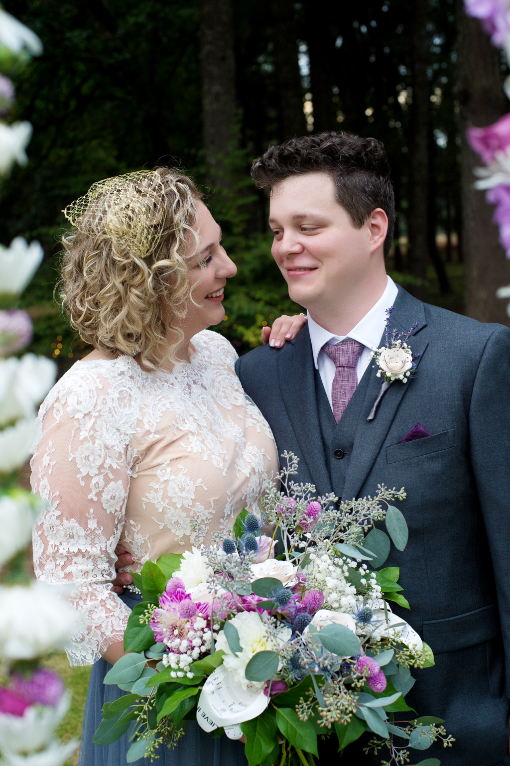 a bride in a lace top with gray flowy skirt holds a pretty purple and ivory wedding bouquet while her groom hugs her