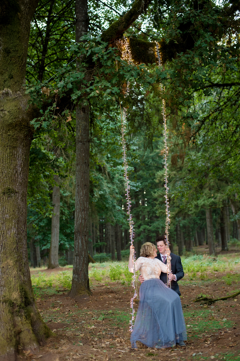 a bride in a flowy gray skirt and ivory lace top swings from a tree swing covered in lights as her groom pushes her