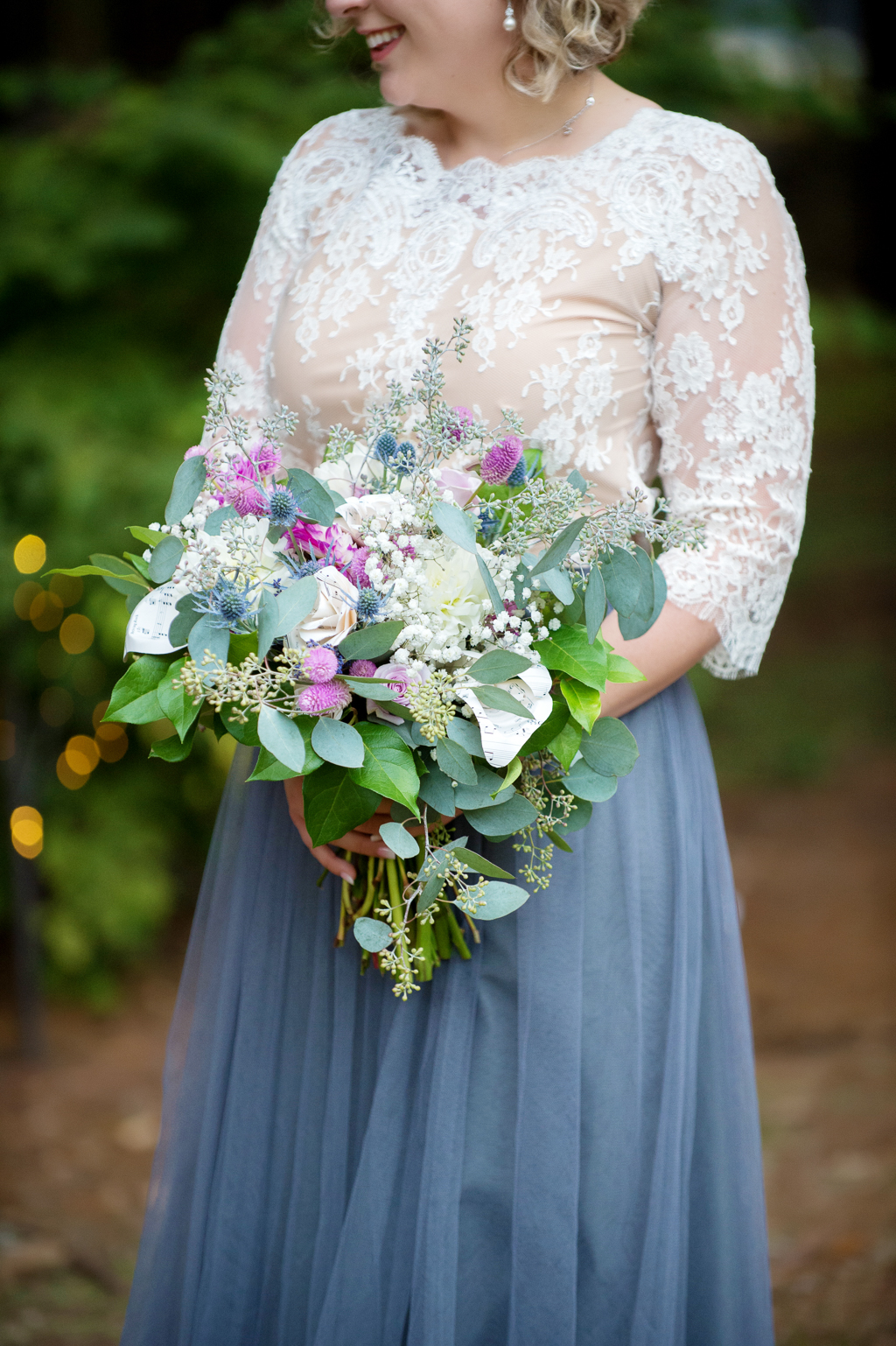 a bride in a lace top with gray flowy skirt holds a pretty purple and ivory wedding bouquet