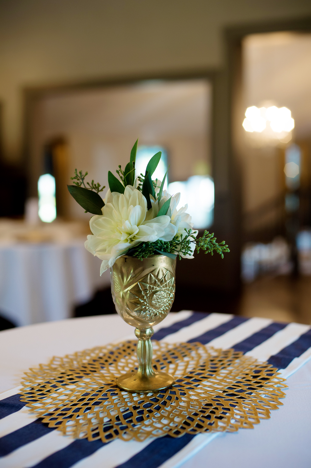 ivory flowers in a gold goblet sit on a gold doily on a navy and blue table runner at a wedding reception 