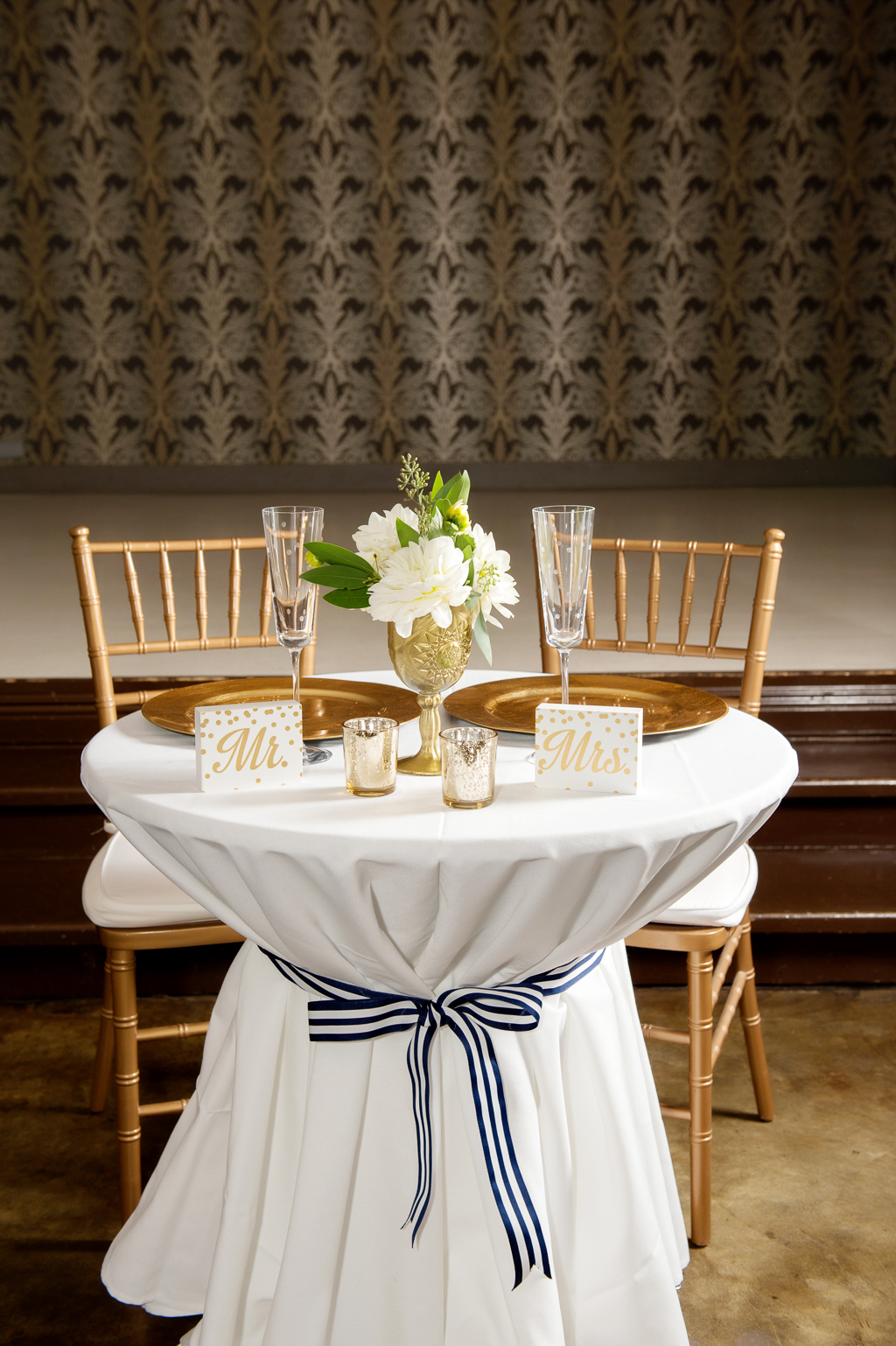 a bride and groom sweetheart table is covered in white cloth and tied with a navy and white ribbon next to gold chairs and a gold goblet filled with ivory flowers