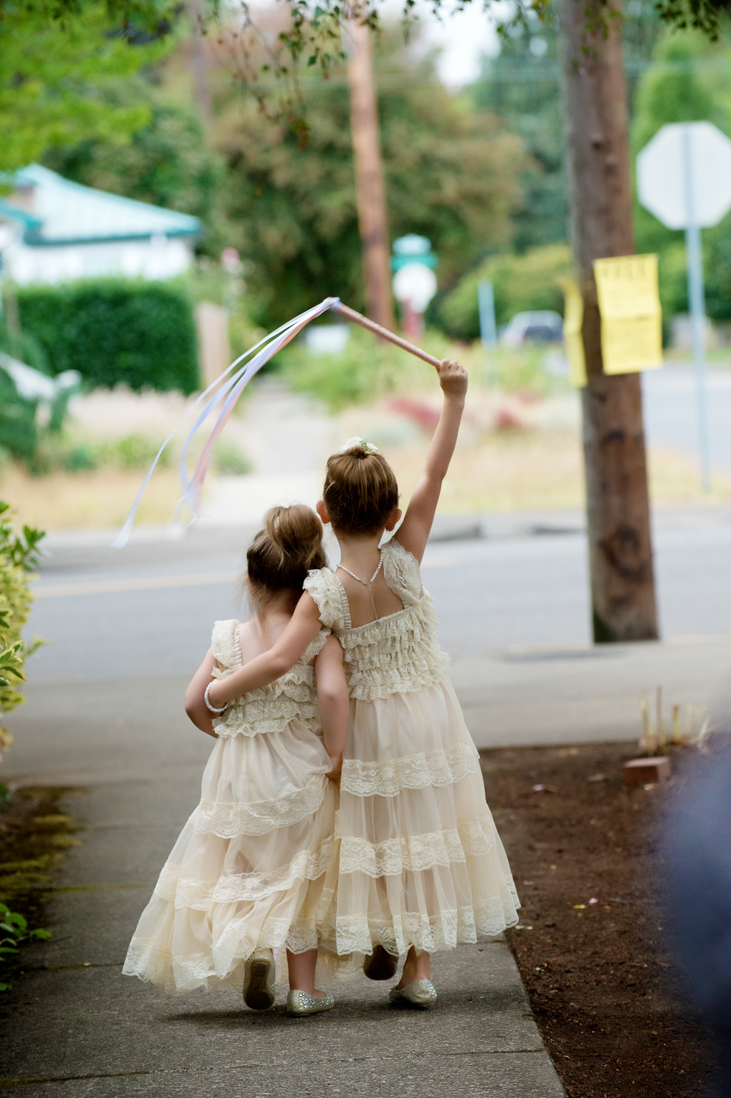 two flower girls walk with their arms around each other waving a ribbon wand in the air