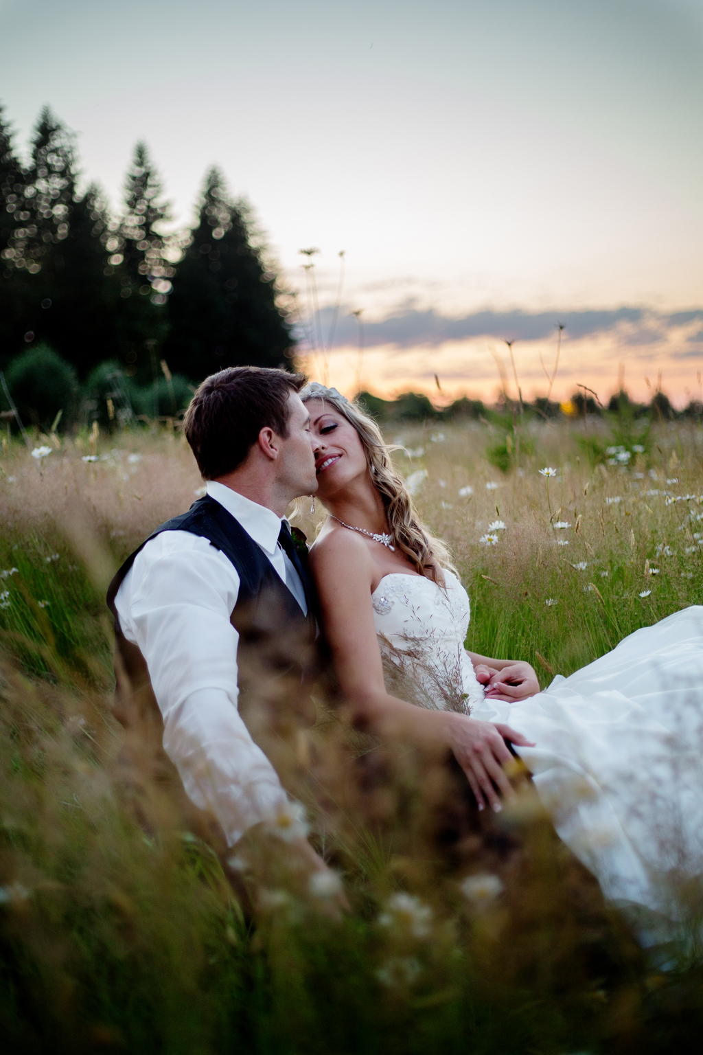 bride and groom sit in tall grass field at sunset