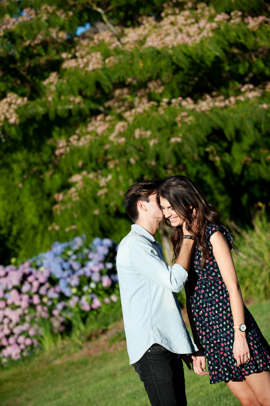 a man whispers something in a girls ear in front of purple hydrangeas and a mimosa tree