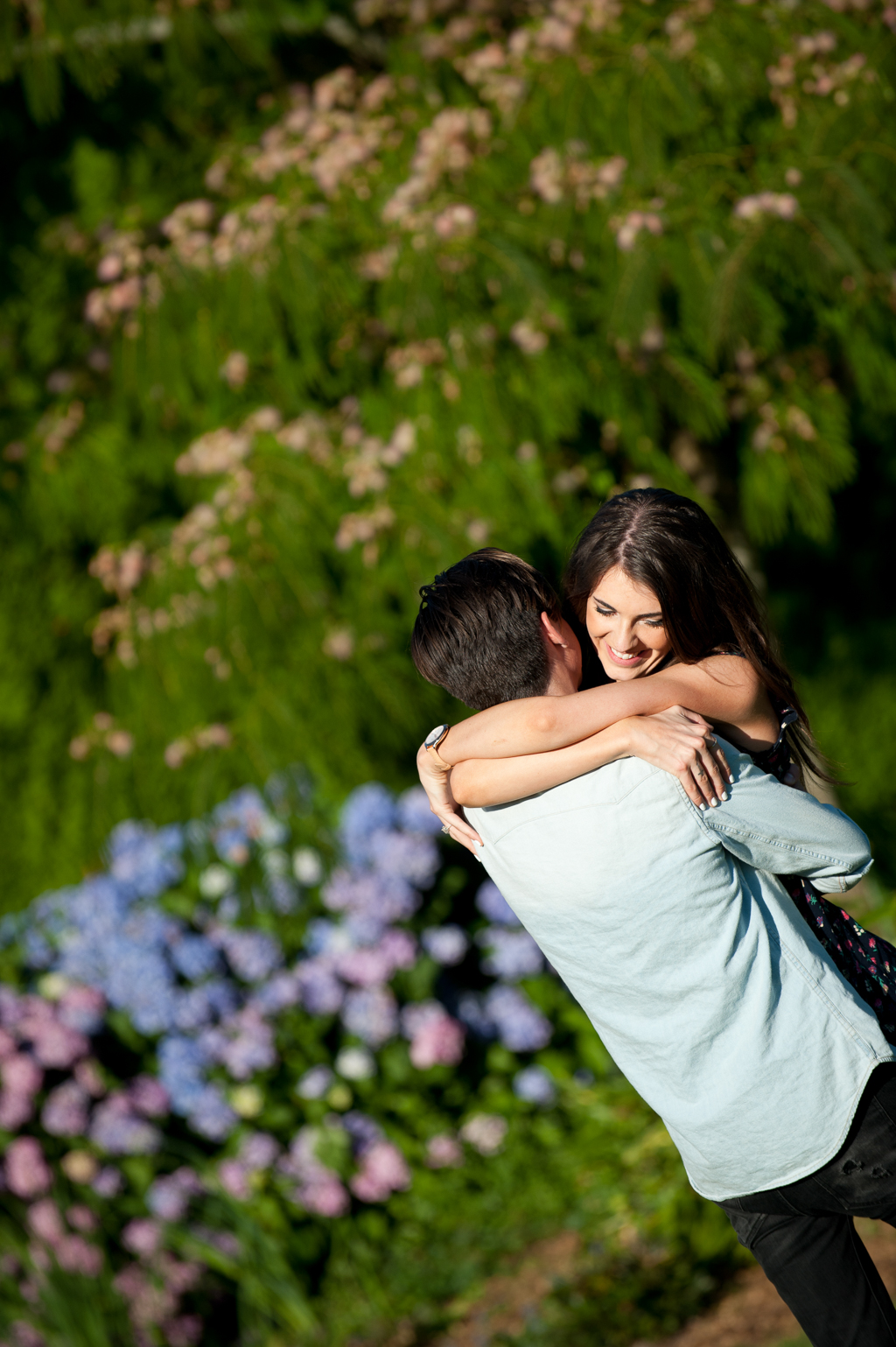 a man picks up a girl and swings her around in front of purple hydrangeas and a mimosa tree