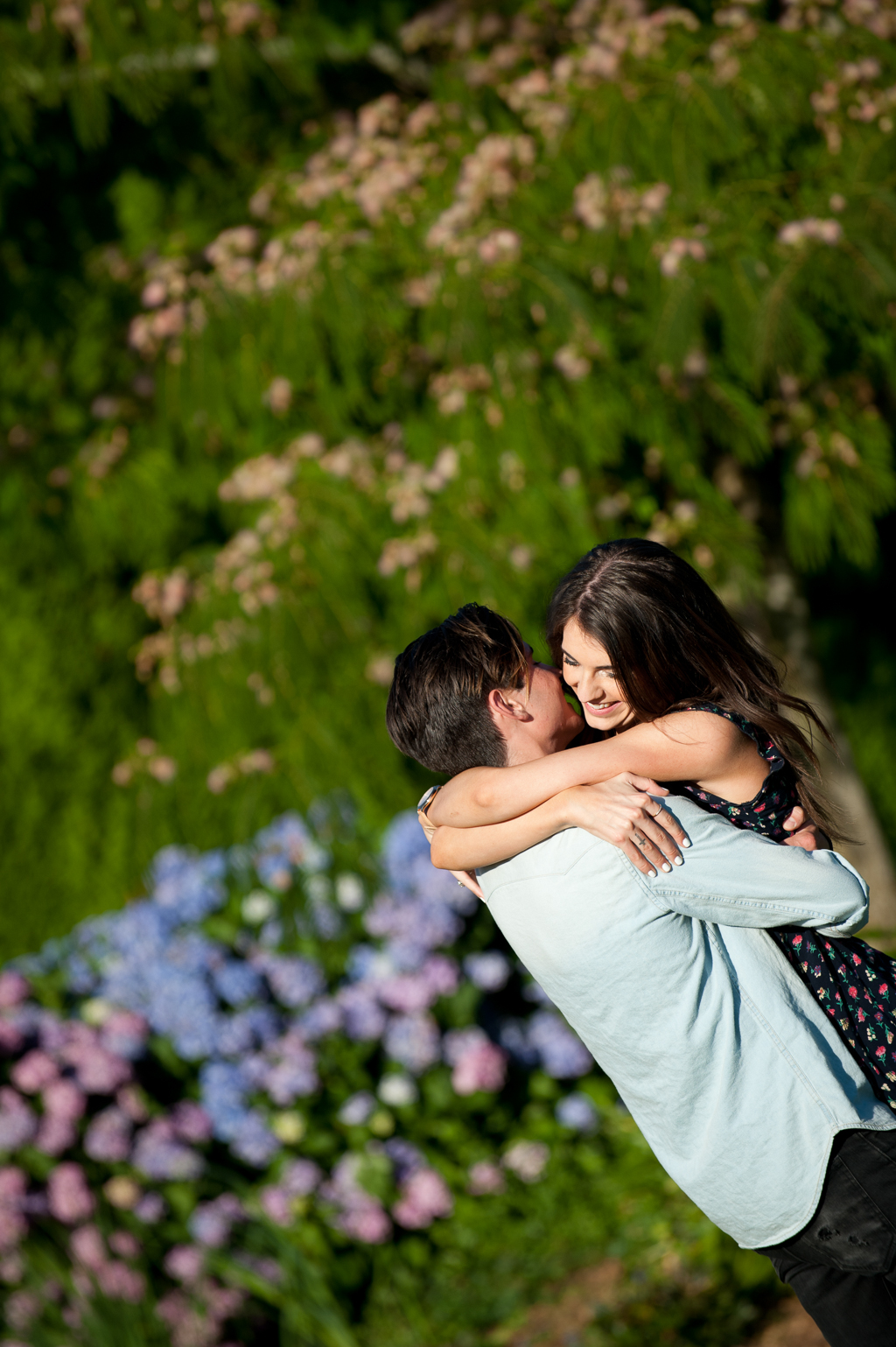 a man picks up a girl and swings her around in front of purple hydrangeas and a mimosa tree