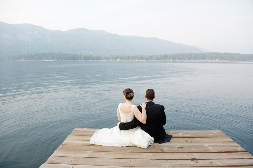 a bride and groom sit on the end of a dock looking out at lake wenatchee on a smoky day that fills the air with fog