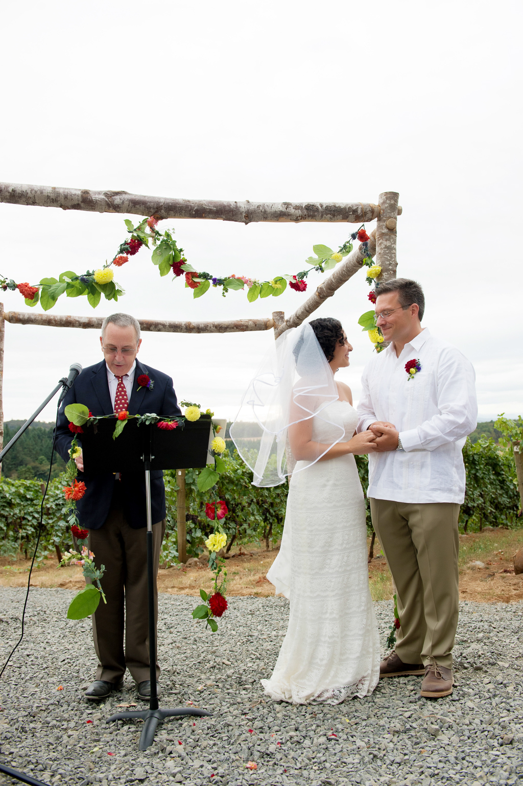 bride and groom stand during wedding ceremony in front of red and yellow flowers hang from a wedding arbor made of birch logs