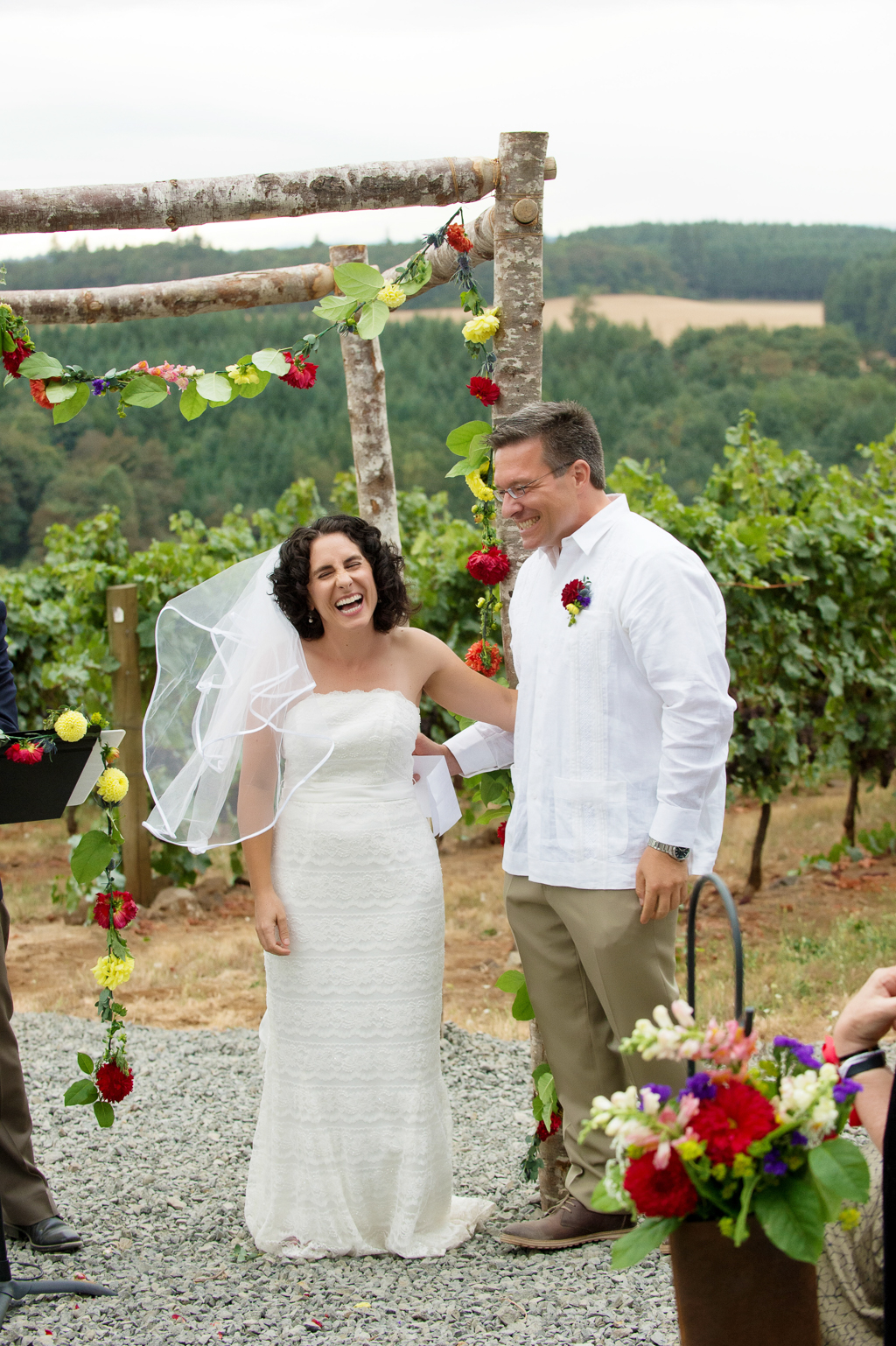 bride and groom stand during wedding ceremony in front of red and yellow flowers hang from a wedding arbor made of birch logs overlooking vineyard