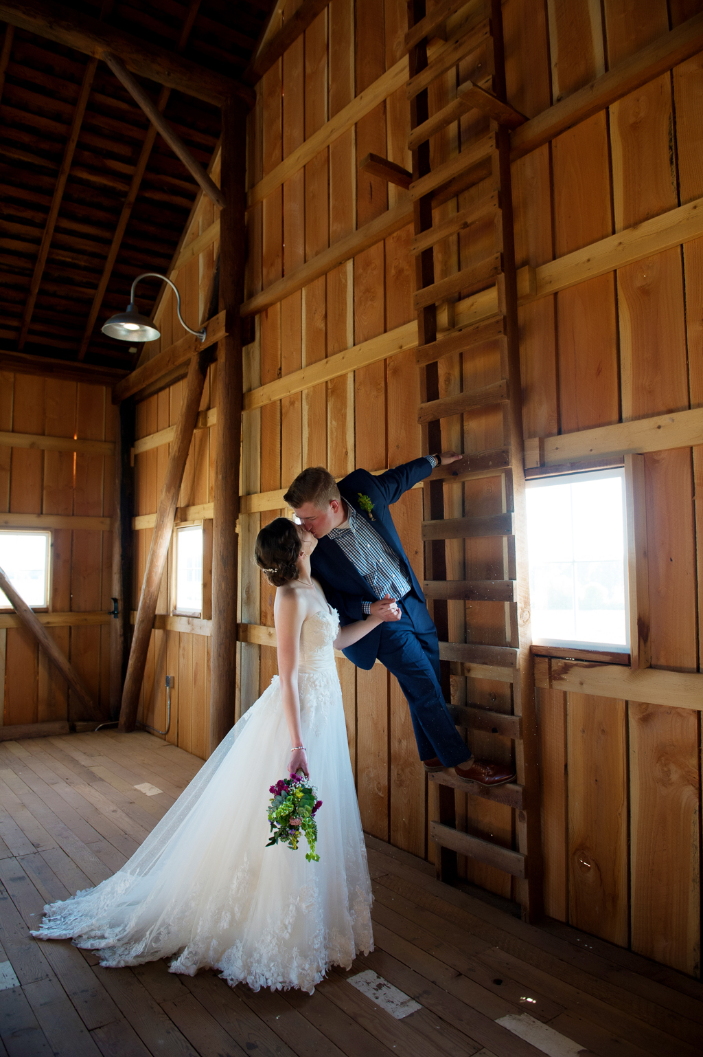groom climbs wooden ladder in barn and leans back to kiss bride