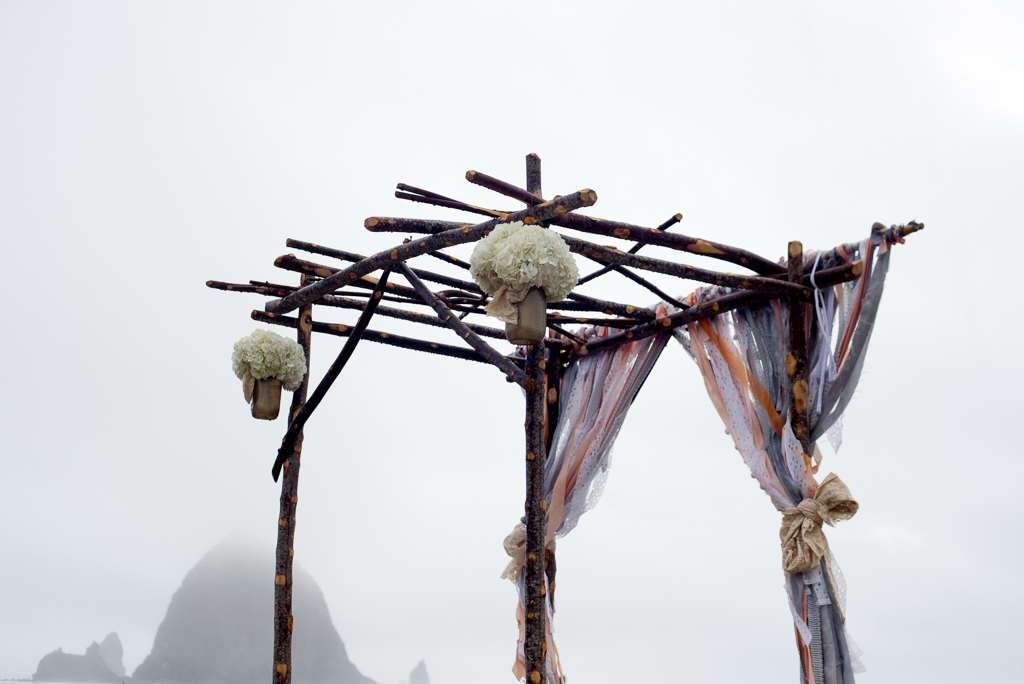 a pretty wedding arbor made of sticks and torn pieces of purple peach and gray cloth with haystack rock in the background