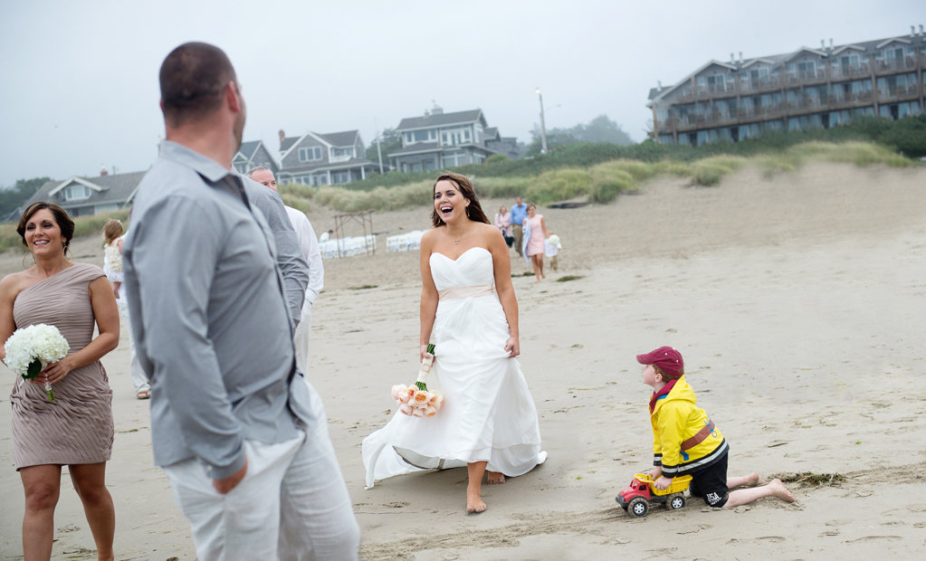 a bride walks barefoot on the sand at the beach while a little boy in a yellow raincoat pushing a dump truck looks up at her as she passes