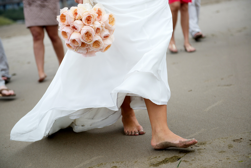 a bride holding a peach bouquet walks barefoot in the sand