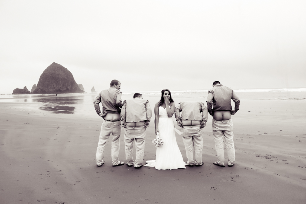 groomsmen stand beside the bride on cannon beach and moon the camera with their butts with haystack rock in the background