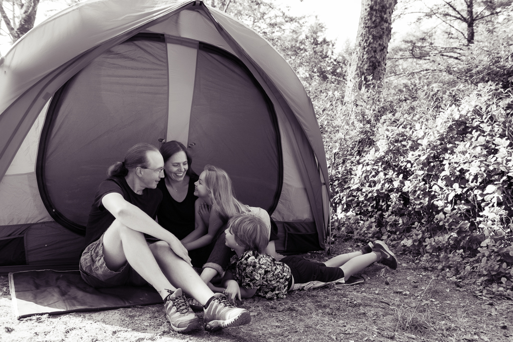 family laughs at each other as they sit on the ground in front of their orange and green tent