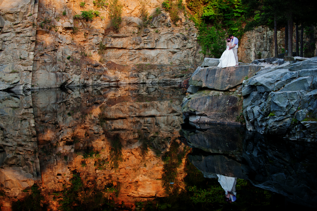 bride and groom kiss on the edge of a rock quarry and their reflection shows in the water