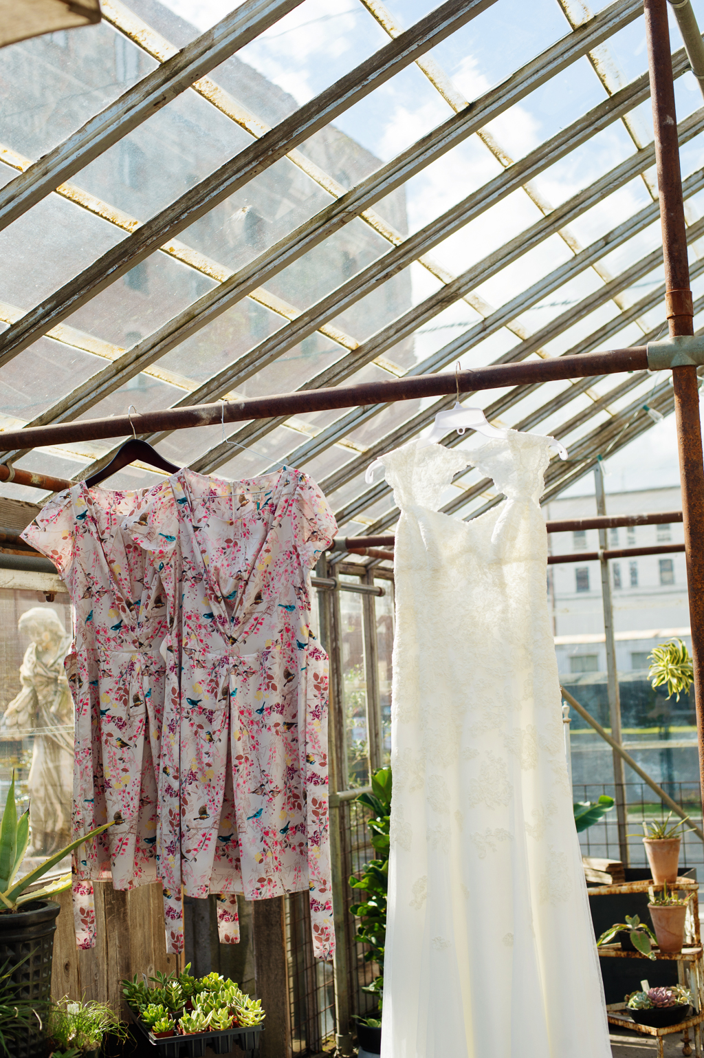 a wedding dress and two pretty multicolored bridesmaid dresses hang from rafters in a glass greenhouse