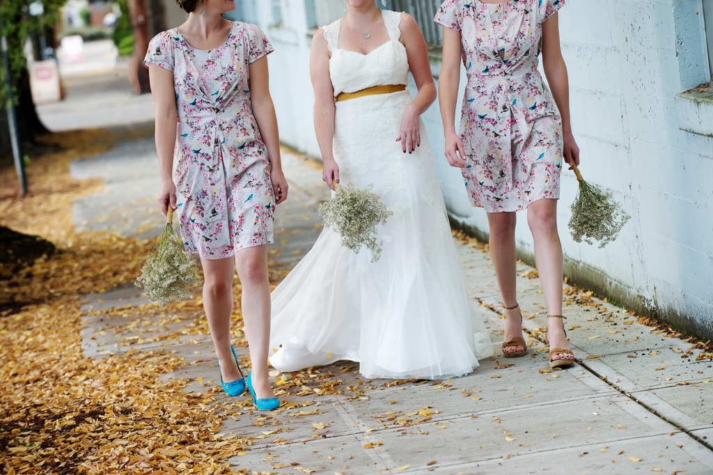 a bride wearing a wedding dress with a yellow mustard ribbon walks between two girls wearing multicolored bird print bridesmaid dresses and turquoise shoes through autumn leaves