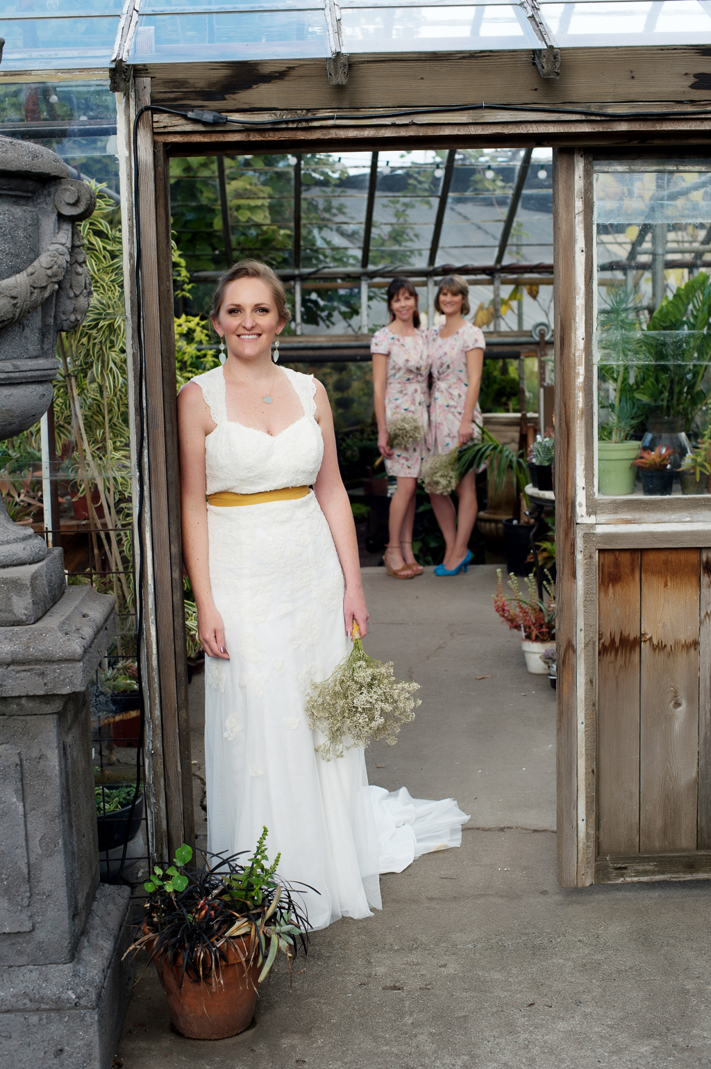 a bride holding a bouquet of baby's breath stands in the doorway of a greenhouse with her bridesmaids in the background