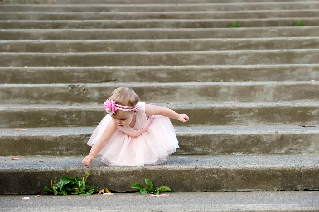 a little girl in a pink dress picks grass growing through the concrete stairs