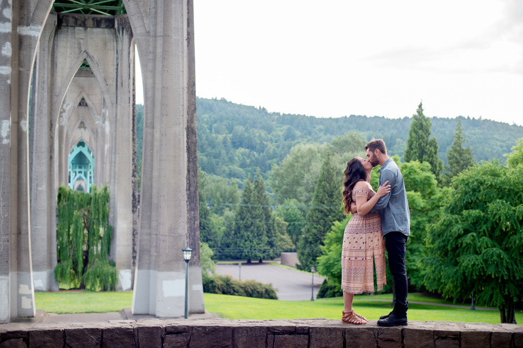 a pregnant woman and a man stand on the wall under the st johns bridge and kiss