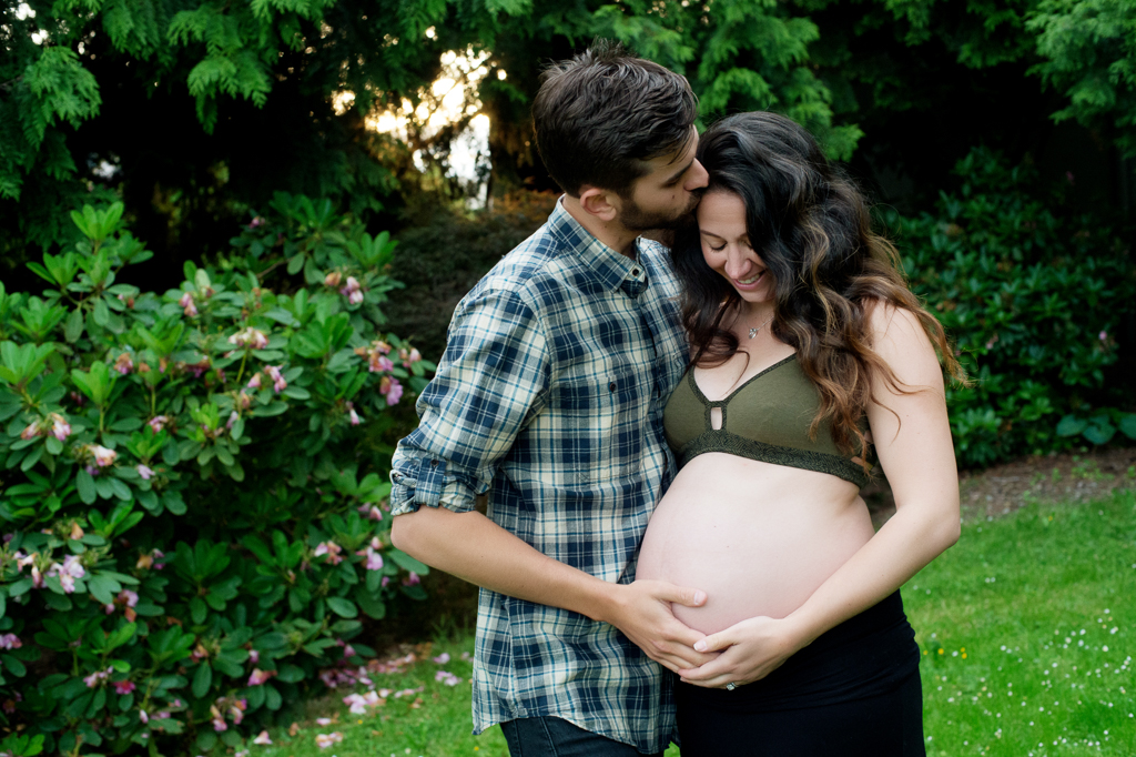 pregnant woman in forest green bralette  looks down at belly as husband kisses her head
