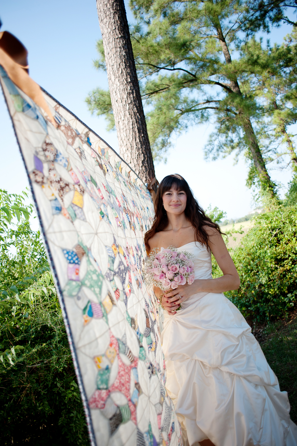a colorful quilt hangs from a tree and a bride stands in front of it holding a bouquet of pink roses and baby's breath