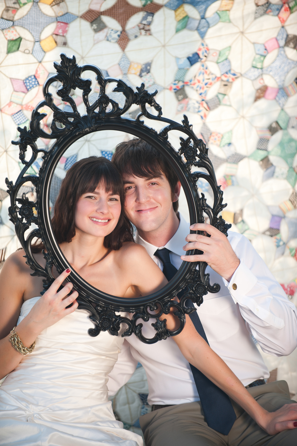 a bride and groom look through an oval photo frame in front of a colorful quilt that hangs from a tree