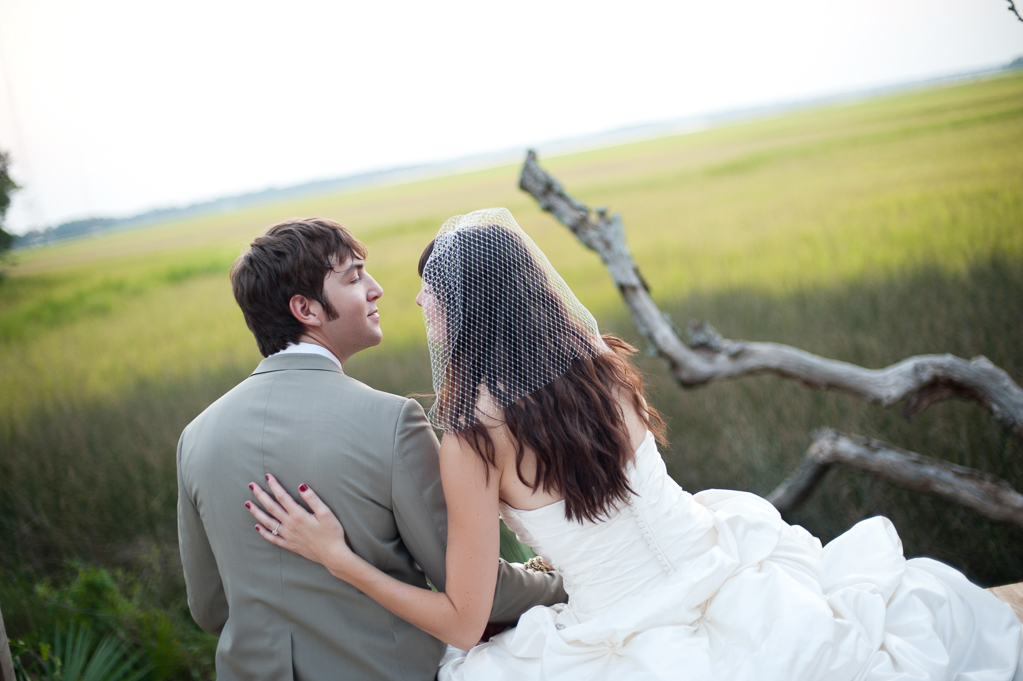 a bride and groom sit overlooking the marsh along the ashley river with a large piece of driftwood tree
