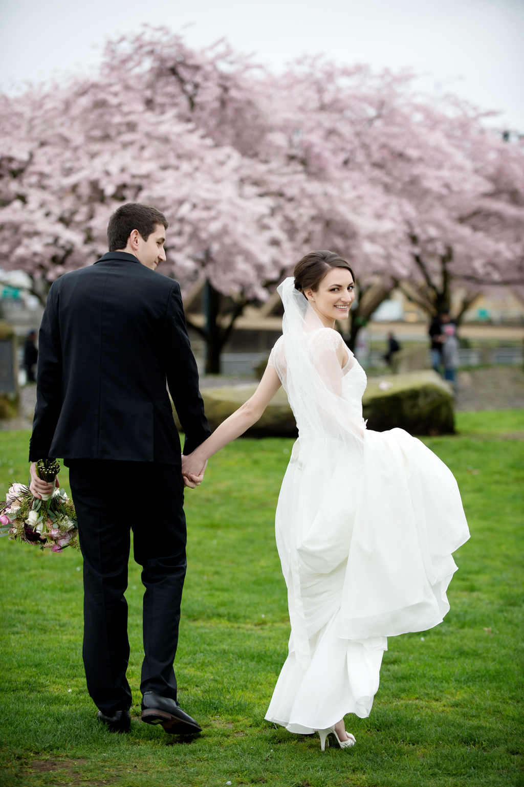 a bride and groom walk holding hands toward cherry blossom trees