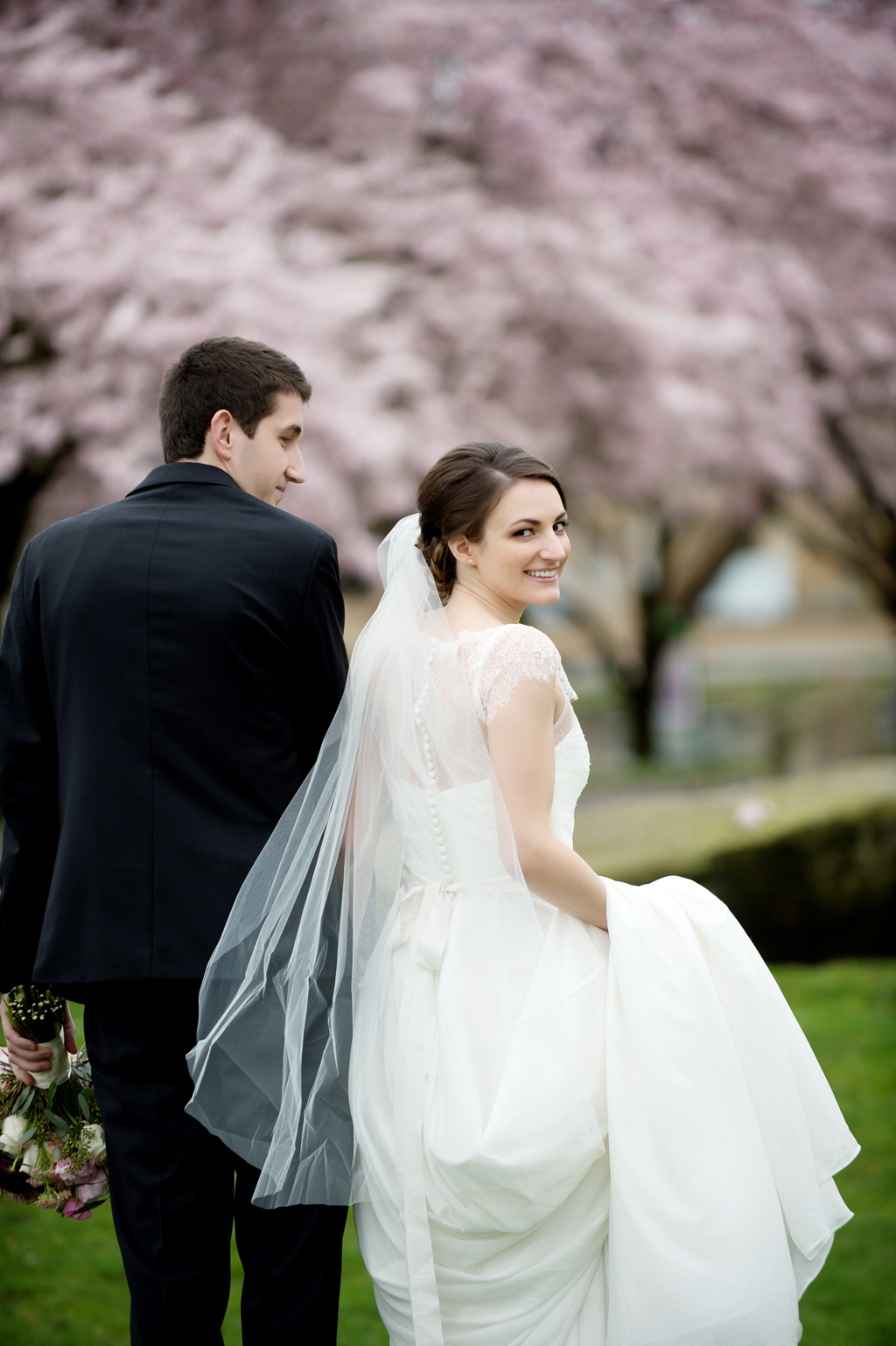a bride looks over her shoulder as she walks with her groom underneath cherry blossom trees